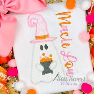 May include: A white t-shirt featuring an embroidered ghost wearing a pink lace-patterned witch hat and holding a dark gray cauldron with orange bubbles. The name Macie Lou is embroidered in orange and pink script next to the ghost design.