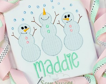 Peut inclure: T-shirt blanc avec une broderie de trois bonhommes de neige avec des nez en carotte et des bras en brindilles. Le nom "Maddie" est brodé en vert en dessous, avec un cœur sur le "i". Des flocons de neige bleus brodés sont dispersés au-dessus.
