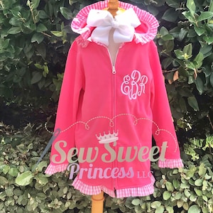 May include: Pink hooded sweatshirt with a white gingham ruffle trim and a white monogram on the front. The sweatshirt has a zipper closure and a white bow on the hood.