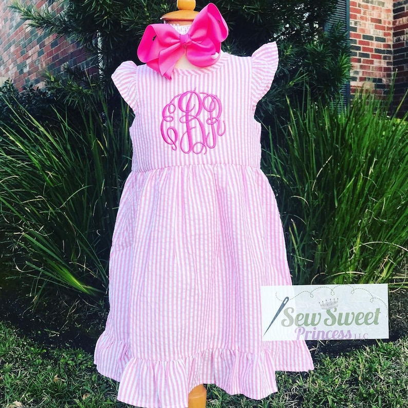 May include: A pink and white striped dress with a pink ruffled neckline and sleeves. The dress has a pink monogram on the front. The dress is on a mannequin and is displayed against a green background.