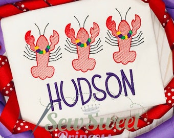 Peut inclure: Un t-shirt blanc avec trois homards rouges brodés portant des colliers de perles violettes et vertes. Le nom "HUDSON" est brodé en violet sous les homards.