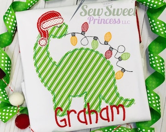 May include: A green and white striped dinosaur wearing a red Santa hat and holding a string of Christmas lights. The dinosaur's name, "Graham", is embroidered below it.
