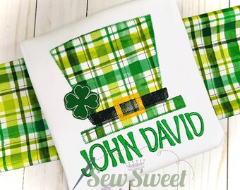 Peut inclure: T-shirt blanc avec un motif de chapeau de lutin à carreaux verts, un trèfle vert et le nom "JOHN DAVID" brodé en vert. Un tissu à carreaux verts et blancs est visible.