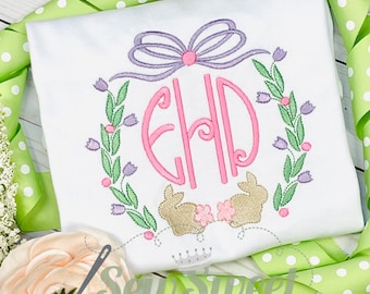 Peut inclure: Un t-shirt blanc avec un motif brodé représentant un monogramme rose "EHD" entouré d'une couronne de feuilles vertes et de fleurs violettes. Deux lapins bruns sont brodés sous le monogramme, avec un nœud rose en haut.