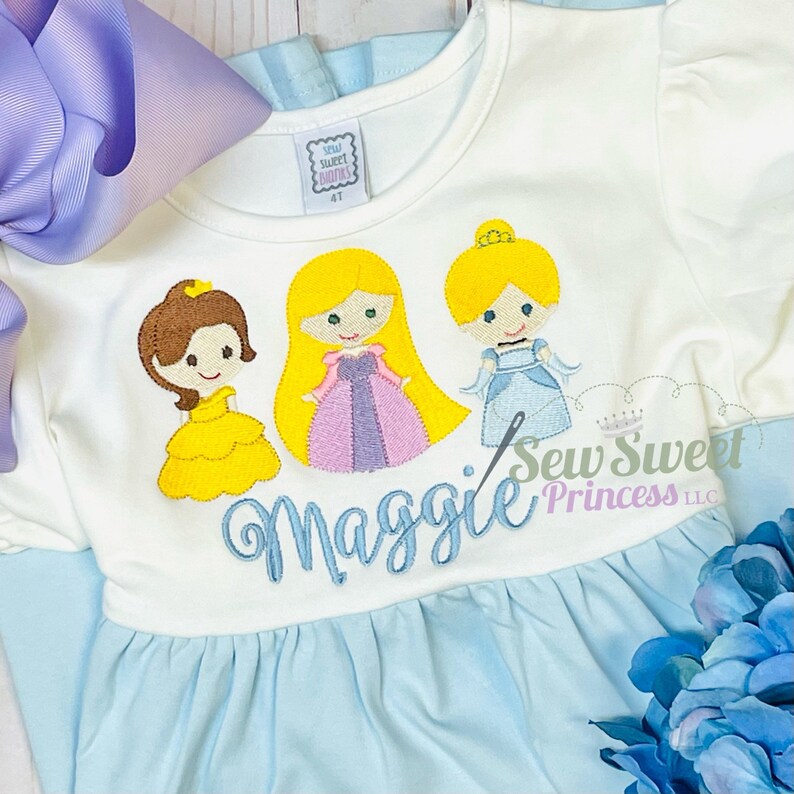 May include: White and light blue dress featuring embroidered cartoon princesses. The dress has a ruffled skirt and sleeves, with the name "Maggie" embroidered in blue script. A large purple bow and blue flowers are also visible.