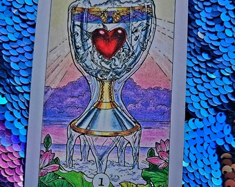 May include: Tarot card depicting the Ace of Cups. The card features a golden chalice filled with water and a red heart floating on top. The chalice is surrounded by a white light and sits on a bed of lotus flowers.