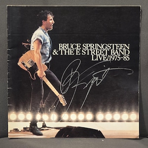 Puede incluir: Una portada de disco de vinilo con Bruce Springsteen tocando una guitarra eléctrica de color claro en el escenario. El texto dice BRUCE SPRINGSTEEN & THE E STREET BAND LIVE/1975-85. Un autógrafo grande en tinta negra está firmado sobre el fondo oscuro.