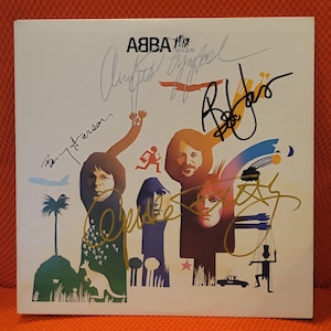 以下が含まれることがあります： ABBA The Albumの正方形のレコードジャケット。4人の人物、ヤシの木、カンガルー、タクシー、マリオネットのカラフルなイラストが描かれています。ジャケットの表面には、バンドメンバーによる黒、金、銀のインクでのサインが入っています。