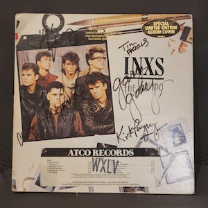 May include: A square vinyl record cover for the INXS album The Swing, featuring a group photograph of the band members, multiple black ink signatures, and a Special Limited Edition Album Cover sticker. The bottom includes an Atco Records tracklist.