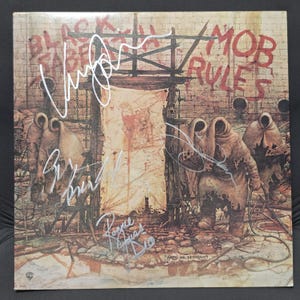 May include: A square vinyl record cover for Black Sabbath Mob Rules featuring a dark, illustrative scene of hooded figures in a stone courtyard with a central wooden structure. The cover includes multiple white ink signatures across the artwork.