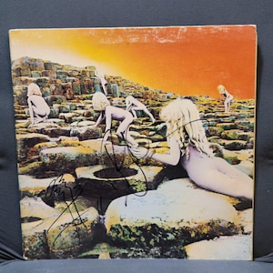 Può includere: Copertina dell'album con un paesaggio con figure che si arrampicano su rocce esagonali sotto un cielo arancione. La copertina ha una firma con inchiostro nero in basso a sinistra. La copertina dell'album è un album rock classico.