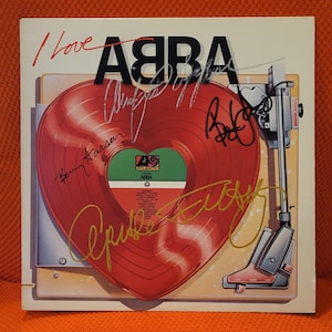 May include: A square vinyl record album cover titled I Love ABBA featuring a large red heart-shaped record graphic and a turntable arm illustration. The cover includes the text ABBA and Atlantic, with multiple handwritten signatures in black and gold ink.