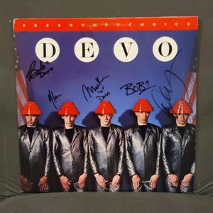 以下が含まれることがあります： デヴォの「フリーダム・オブ・チョイス」のアルバムカバー。カバーには、銀色のジャンプスーツと赤い帽子をかぶった5人のバンドメンバーが写っており、バンド名は青い背景に白い円で表示されています。アルバムカバーにはサインが入っています。