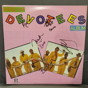 以下が含まれることがあります： ピンク色のアルバムカバー。タイトル「DEVOTEES」がカラフルなブロックレターで表示されています。黄色いつなぎを着たバンドの写真とサインが特徴です。「ALBUM」というラベルが青いボックスに表示されています。