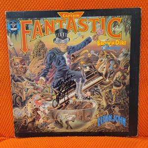 Könnte beinhalten: Albumcover für "Captain Fantastic and the Brown Dirt Cowboy" von Elton John. Das Artwork zeigt eine cartoonartige Illustration eines Mannes mit Zylinder und Maske, mit einem Klavier und verschiedenen fantastischen Kreaturen. Das Albumcover ist signiert.