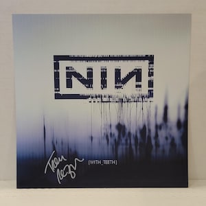 May include: A square vinyl record cover featuring the Nine Inch Nails logo in a dark blue, glitch-style rectangular frame. Below the logo, the text [WITH_TEETH] appears in white, accompanied by a handwritten signature in white ink on the left side.