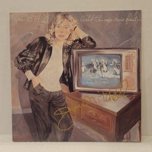 Könnte beinhalten: Ein Schallplattencover mit einem Gemälde einer Person mit blondem Haar, die eine schwarze Jacke und ein weißes Hemd trägt und sich an einen Fernseher lehnt, auf dem Pferde im Wasser laufen. Der Text Joni Mitchell Wild things run fast ist zu sehen.