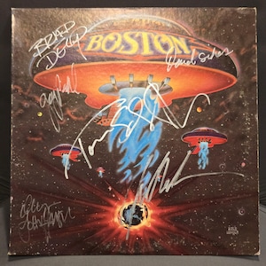Pode incluir: Uma capa de disco de vinil quadrada da banda Boston com uma ilustração de tema espacial de discos voadores laranja emitindo luz azul sobre um planeta, assinada por vários membros da banda com uma caneta de cor prateada.