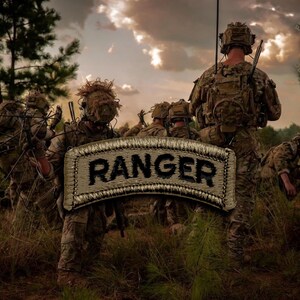 Pode incluir: Um emblema retangular bordado com fundo bege e borda preta apresentando a palavra RANGER em letras maiúsculas pretas em negrito, sobreposto a uma cena de soldados em uniformes de camuflagem e equipamento tático em um campo de grama.