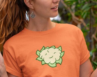 May include: A woman wearing an orange t-shirt with a cartoon cauliflower graphic. The shirt has a knotted front and the woman is wearing a blue skirt.