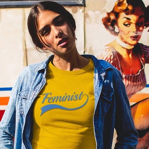 Puede incluir: Una mujer lleva una camiseta amarilla con la palabra "Feminista" en letras azules sobre un gr&aacute;fico de estilo vintage. Lleva una chaqueta vaquera y unos vaqueros rotos. El fondo es un mural de estilo vintage de una mujer.