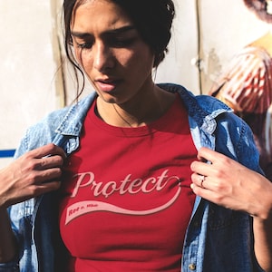 May include: A woman wearing a red t-shirt with the text "Protect More Than This" in a pink cursive font. She is wearing a blue denim jacket over the shirt.