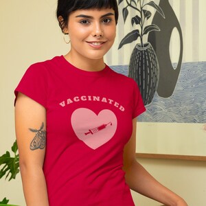 May include: A red t-shirt with a pink heart and a syringe graphic. The text "VACCINATED" is printed in white letters above the heart.