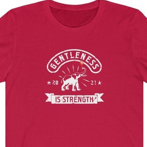 Puede incluir: Una camiseta roja con un gr&aacute;fico blanco. El gr&aacute;fico incluye la palabra "GENTLENESS" en un dise&ntilde;o arqueado sobre una ilustraci&oacute;n de un elefante. Debajo del elefante est&aacute; la frase "IS STRENGTH" en una pancarta. La camiseta tambi&eacute;n tiene los n&uacute;meros "20" y "21" con estrellas.