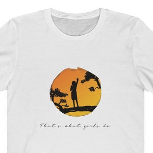 Puede incluir: Camiseta blanca con la silueta de una persona de pie en una rama de un &aacute;rbol frente a una puesta de sol. El texto "That's what girls do" est&aacute; impreso debajo de la imagen.