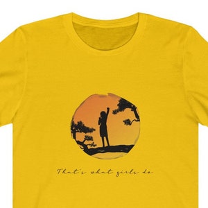 Puede incluir: Camiseta amarilla con una silueta de una persona de pie en una colina con un &aacute;rbol frente a una puesta de sol. El texto "That's what girls do" est&aacute; impreso debajo de la imagen.