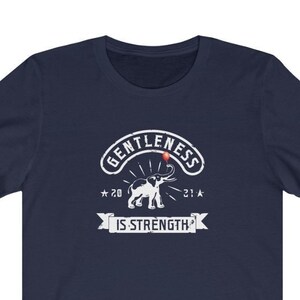 Puede incluir: Camiseta azul marino con un gr&aacute;fico blanco. El gr&aacute;fico presenta las palabras "GENTLENESS IS STRENGTH" con una ilustraci&oacute;n de elefante. El dise&ntilde;o incluye los a&ntilde;os "20" y "21" con acentos de estrella.