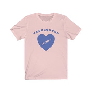 May include: A pink t-shirt with the text "VACCINATED" in blue letters and a blue heart with a syringe inside.