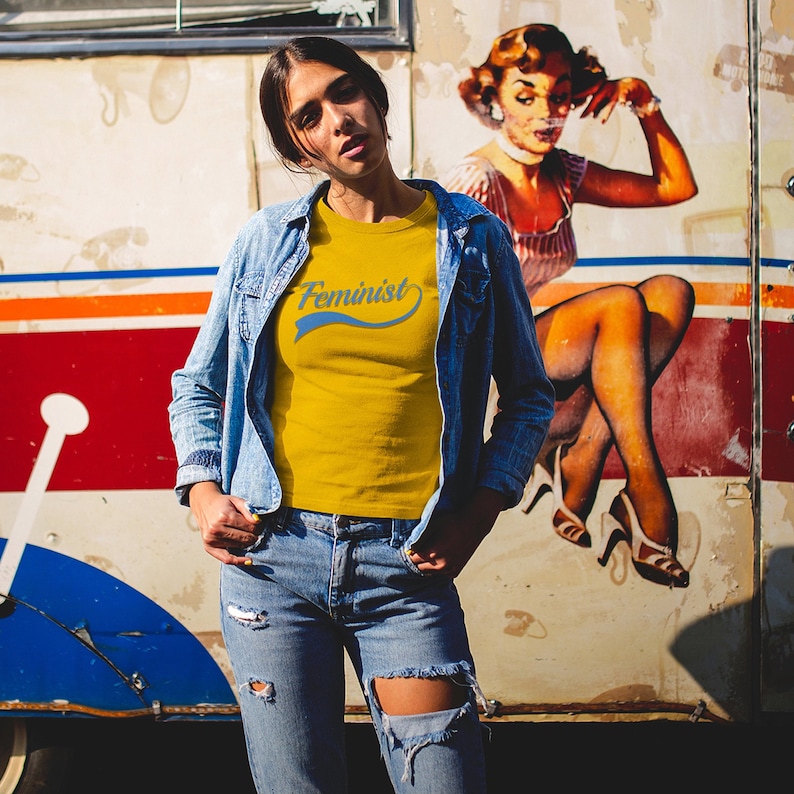 Puede incluir: Una mujer lleva una camiseta amarilla con la palabra "Feminista" en letras azules sobre un gr&aacute;fico de estilo vintage. Lleva una chaqueta vaquera y unos vaqueros rotos. El fondo es un mural de estilo vintage de una mujer.