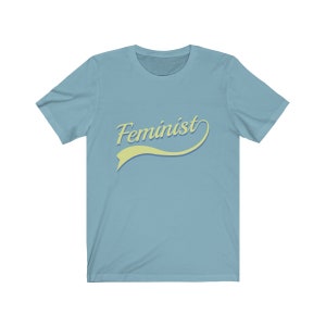 Puede incluir: Una camiseta azul claro con la palabra "Feminista" en una fuente retro amarilla y azul.