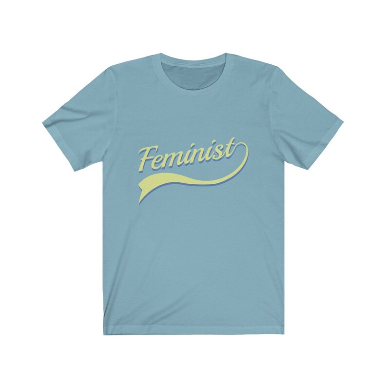 Puede incluir: Una camiseta azul claro con la palabra "Feminista" en una fuente retro amarilla y azul.