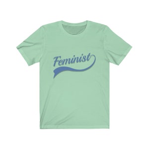 Puede incluir: Una camiseta verde claro con un gr&aacute;fico azul y blanco que dice "Feminista" en una fuente de estilo retro.