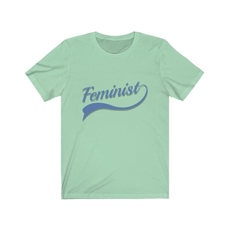 Puede incluir: Una camiseta verde claro con un gr&aacute;fico azul y blanco que dice "Feminista" en una fuente de estilo retro.