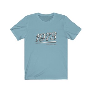 May include: Light blue t-shirt with a silver graphic of the year 1973 and the text "Positive vibes only".