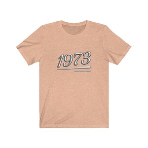 May include: A light peach colored t-shirt with a silver graphic of the year 1973 with a silver line underneath. The text "PAST YEARS DON'T COUNT" is written in small letters underneath the year.