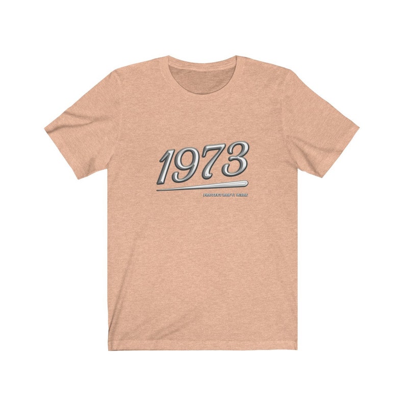 May include: A light peach colored t-shirt with a silver graphic of the year 1973 with a silver line underneath. The text "PAST YEARS DON'T COUNT" is written in small letters underneath the year.