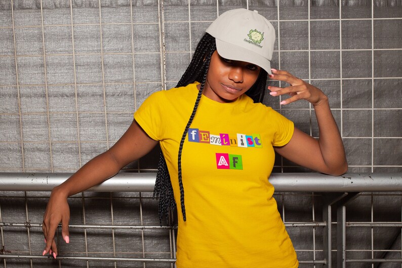 May include: A woman wearing a yellow t-shirt with the text "feminist AF" in colorful block letters. She is wearing a beige baseball cap with a green cartoon logo.