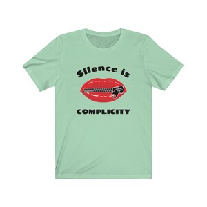 Puede incluir: Una camiseta verde claro con un gr&aacute;fico negro de una boca roja con una cremallera subida. El texto "Silence is Complicity" est&aacute; impreso debajo del gr&aacute;fico.