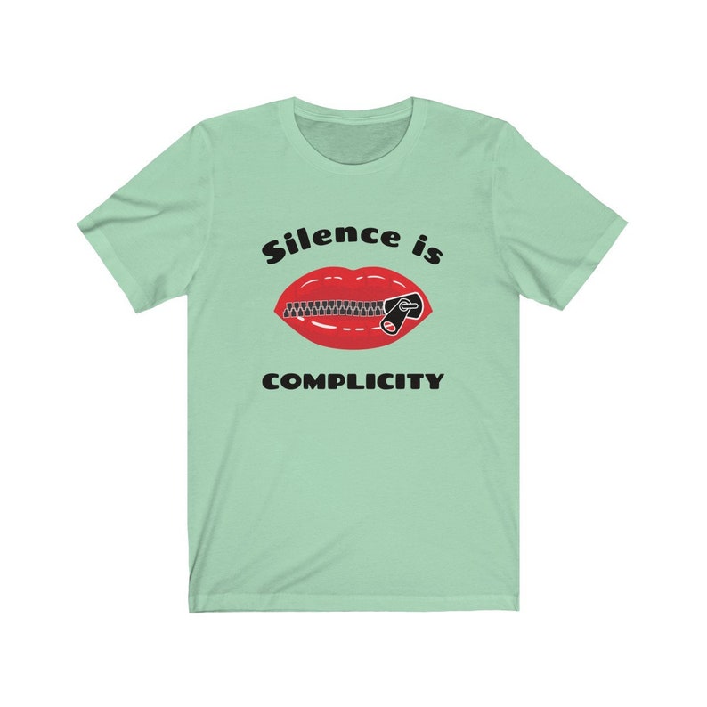 Puede incluir: Una camiseta verde claro con un gr&aacute;fico negro de una boca roja con una cremallera subida. El texto "Silence is Complicity" est&aacute; impreso debajo del gr&aacute;fico.