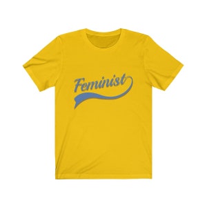 Puede incluir: Camiseta amarilla con la palabra "Feminista" en una fuente azul cursiva con un swoosh azul debajo.