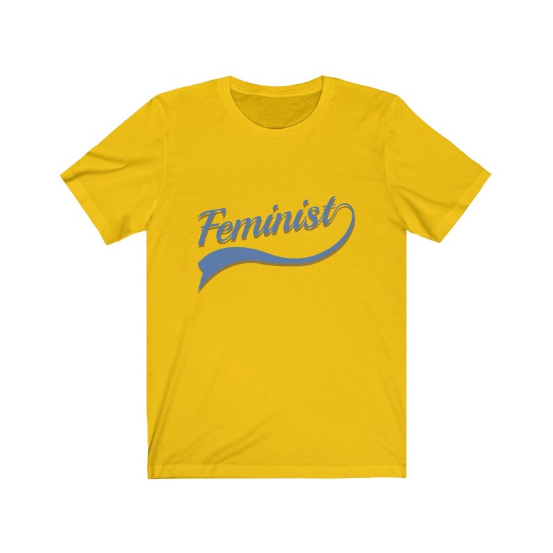 Puede incluir: Camiseta amarilla con la palabra "Feminista" en una fuente azul cursiva con un swoosh azul debajo.