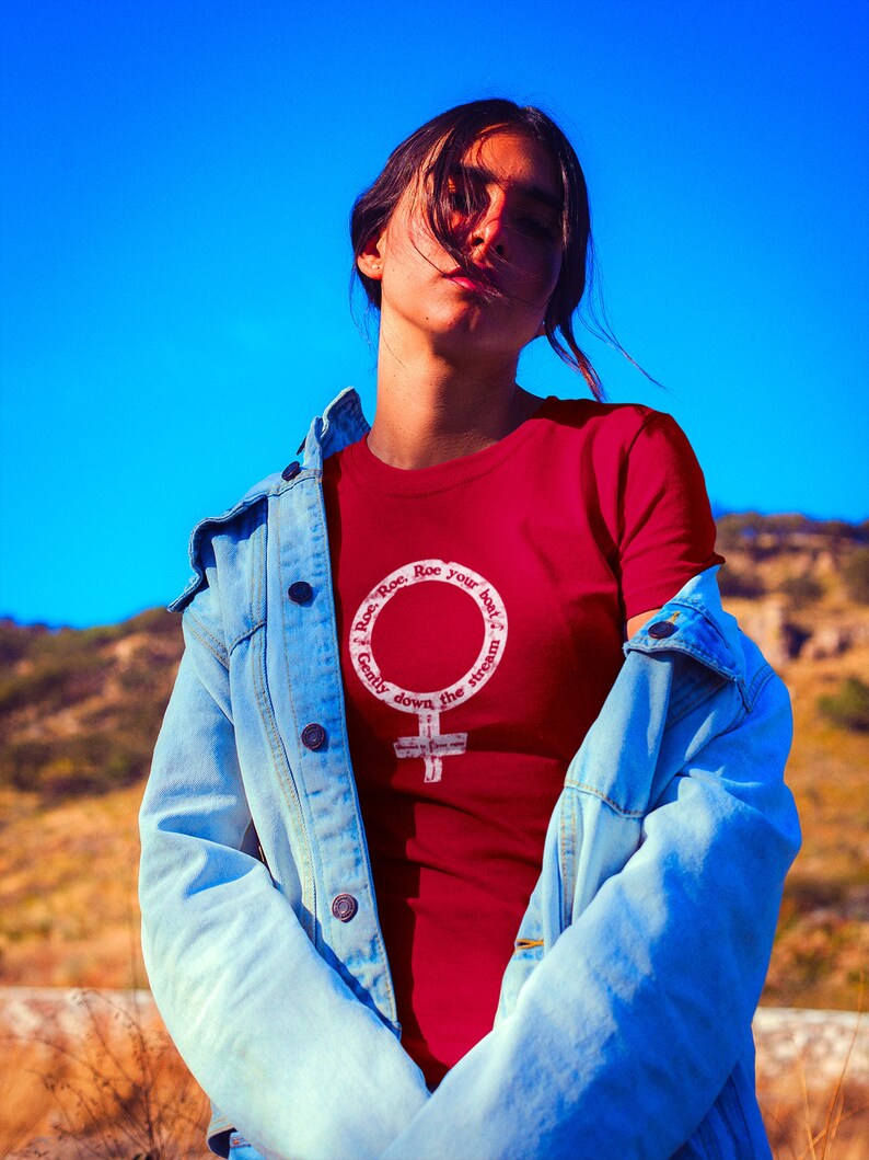 May include: A woman wearing a red t-shirt with a white feminist symbol and the text "Roe. Roe. Roe your boat. Gently down the stream." The woman is wearing a light blue denim jacket.