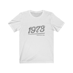 May include: White t-shirt with a silver 1973 graphic and the text "Unapologetically 1973" underneath.