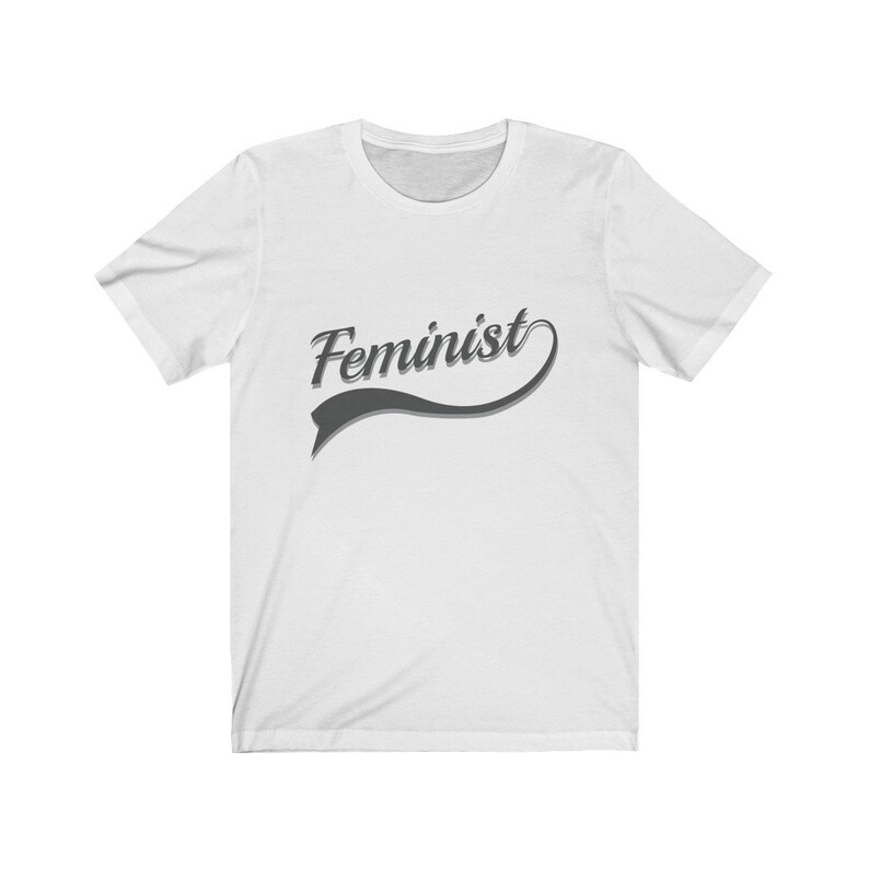 Puede incluir: Camiseta blanca con un gr&aacute;fico gris que dice "Feminista" en una fuente de estilo retro.