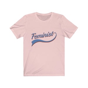 Puede incluir: Una camiseta rosa con la palabra "Feminista" en una fuente retro azul y roja.