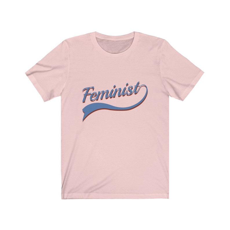 Puede incluir: Una camiseta rosa con la palabra "Feminista" en una fuente retro azul y roja.